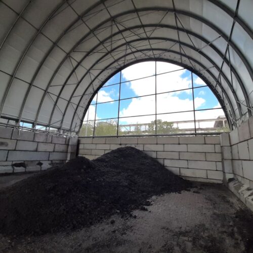 Na zdjęciu widać wnętrze hali namiotowej z bloków betonowych i poszyciem z membrany PCV, w której składowany jest materiał o ciemnej barwie - prawdopodobnie frakcja RDF. Hala ma konstrukcję łukową, wspartą na stalowym szkielecie, z wysokimi ścianami oporowymi z bloczków betonowych.