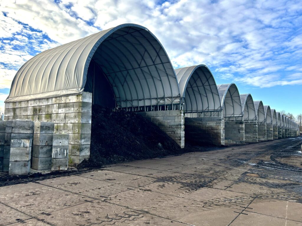 Na zdjęciu widzimy rząd dużych hal łukowych przeznaczonych do składowania osadów lub materiałów sypkich. Konstrukcje wykonane są ze stalowych łuków, obudowanych membraną PCV w jasnoszarym kolorze. Ściany boczne zbudowane zostały z betonowych bloków, które zabezpieczają składowany materiał przed osuwaniem się. Pod jedną z hal widoczna jest wysoka hałda ciemnego osadu. Całość znajduje się na utwardzonym placu, a niebo nad halami pokryte jest częściowo chmurami.