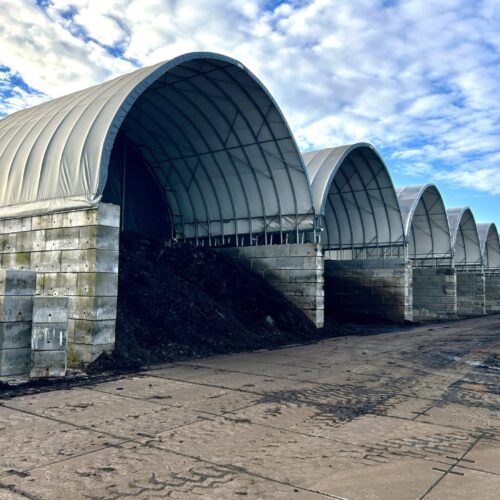 Na zdjęciu widzimy rząd dużych hal łukowych przeznaczonych do składowania osadów lub materiałów sypkich. Konstrukcje wykonane są ze stalowych łuków, obudowanych membraną PCV w jasnoszarym kolorze. Ściany boczne zbudowane zostały z betonowych bloków, które zabezpieczają składowany materiał przed osuwaniem się. Pod jedną z hal widoczna jest wysoka hałda ciemnego osadu. Całość znajduje się na utwardzonym placu, a niebo nad halami pokryte jest częściowo chmurami.