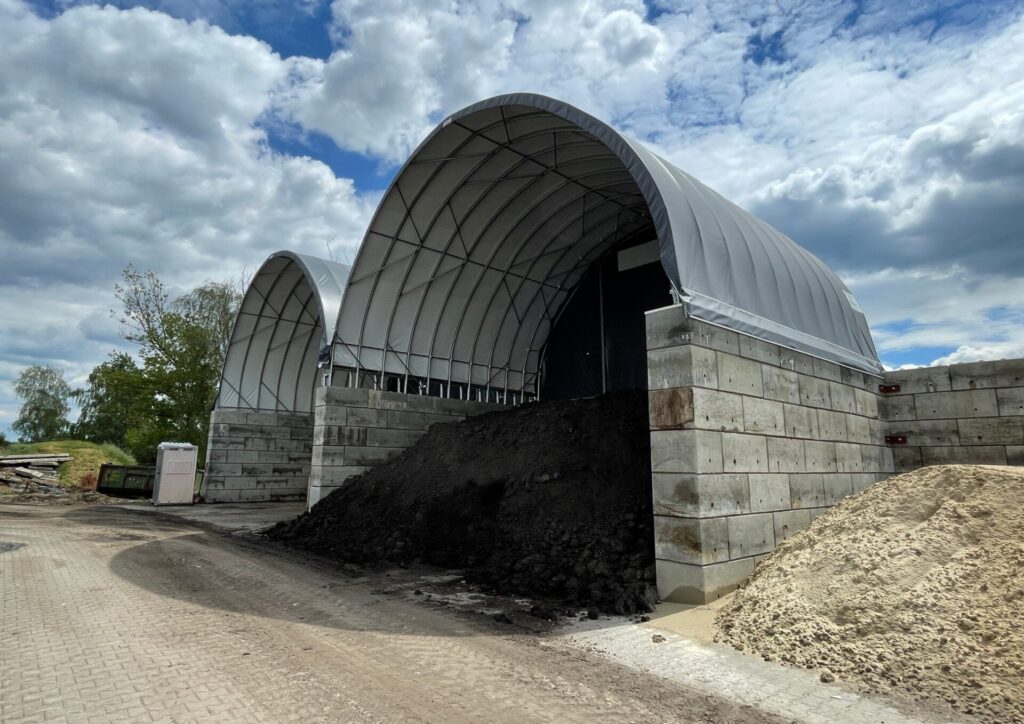 Na zdjęciu widzimy dwie hale łukowe przeznaczone do składowania osadów. Konstrukcje zbudowane są z betonowych bloków tworzących ściany oporowe oraz stalowych łuków przykrytych jasną membraną PCV. Pod zadaszeniem znajduje się duża pryzma ciemnego osadu, a obok niej hałda jasnego kruszywa. Dach hali chroni materiały przed opadami atmosferycznymi i wiatrem.