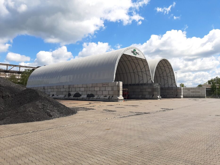 Na zdjęciu widzimy halę namiotową z łukowym dachem, przeznaczoną do składowania i zabezpieczania frakcji paliw alternatywnych RDF (Refuse-Derived Fuel). Konstrukcja opiera się na stalowych ramach z poszyciem z membrany PCV, a ściany zbudowane są z betonowych bloków, które zapewniają stabilność i oddzielenie materiału.