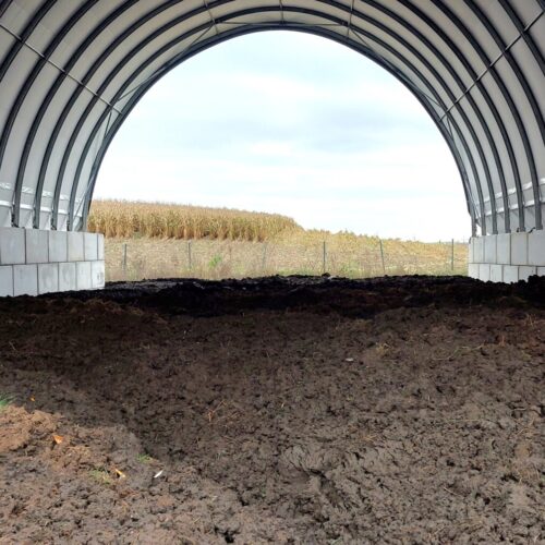 Na zdjęciu widzimy halę łukową wykorzystywaną jako zadaszenie dla kompostu. Wewnątrz obiektu zgromadzona jest duża ilość ciemnej masy organicznej, stanowiącej materiał w trakcie procesu kompostowania. Ściany hali zbudowane są z betonowych bloków, a dach wykonany z jasnej membrany PCV rozpiętej na stalowej konstrukcji łukowej. Otwarta przestrzeń umożliwia wentylację, a jednocześnie chroni składowany materiał przed opadami i bezpośrednim nasłonecznieniem.