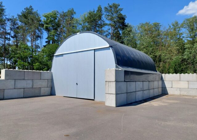 Na zdjęciu widzimy halę łukową pełniącą funkcję zadaszenia kompostowni. Konstrukcja została wykonana ze stalowych łuków, a dach pokryto membraną PCV w ciemnoszarym kolorze. Front obiektu zamknięty jest dużymi, przesuwnymi drzwiami z blachy trapezowej, które umożliwiają łatwy dostęp do wnętrza. Ściany boczne i tylne zbudowane są z betonowych bloków, które stabilizują całość i chronią materiał organiczny przed wysypywaniem się na zewnątrz.