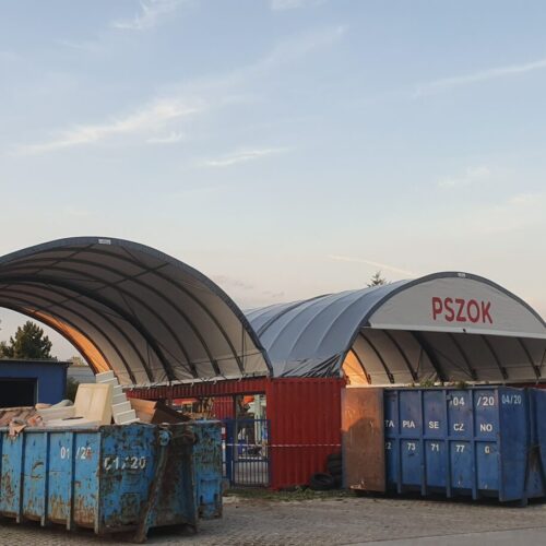 Na zdjęciu widzimy zadaszone wiaty łukowe z napisem „PSZOK”, czyli Punkt Selektywnej Zbiórki Odpadów Komunalnych. Konstrukcje oparte są na stalowym stelażu i przykryte membraną PCV, co chroni składowane materiały przed warunkami atmosferycznymi. Przed halami ustawiono duże metalowe kontenery na różne frakcje odpadów, m.in. gabaryty, tworzywa czy złom. Całość stanowi zorganizowaną przestrzeń do gromadzenia i segregacji odpadów dostarczanych przez mieszkańców.