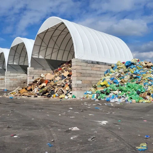 Na zdjęciu widać rząd zadaszonych hal łukowych z białą membraną PCV, pełniących funkcję boksów do magazynowania odpadów.