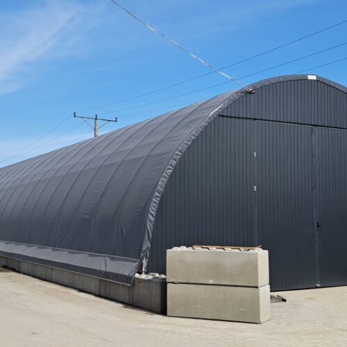 Zamknięta hala namiotowa o konstrukcji łukowej, pokryta ciemnoszarą plandeką i wyposażona w front z metalowych paneli, ustawiona na betonowych blokach.