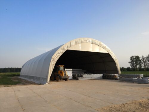 Duża hala namiotowa w jasnym kolorze, wykorzystywana jako magazyn zewnętrzny, z widocznym w środku sprzętem budowlanym i stosami metalowych elementów konstrukcyjnych.