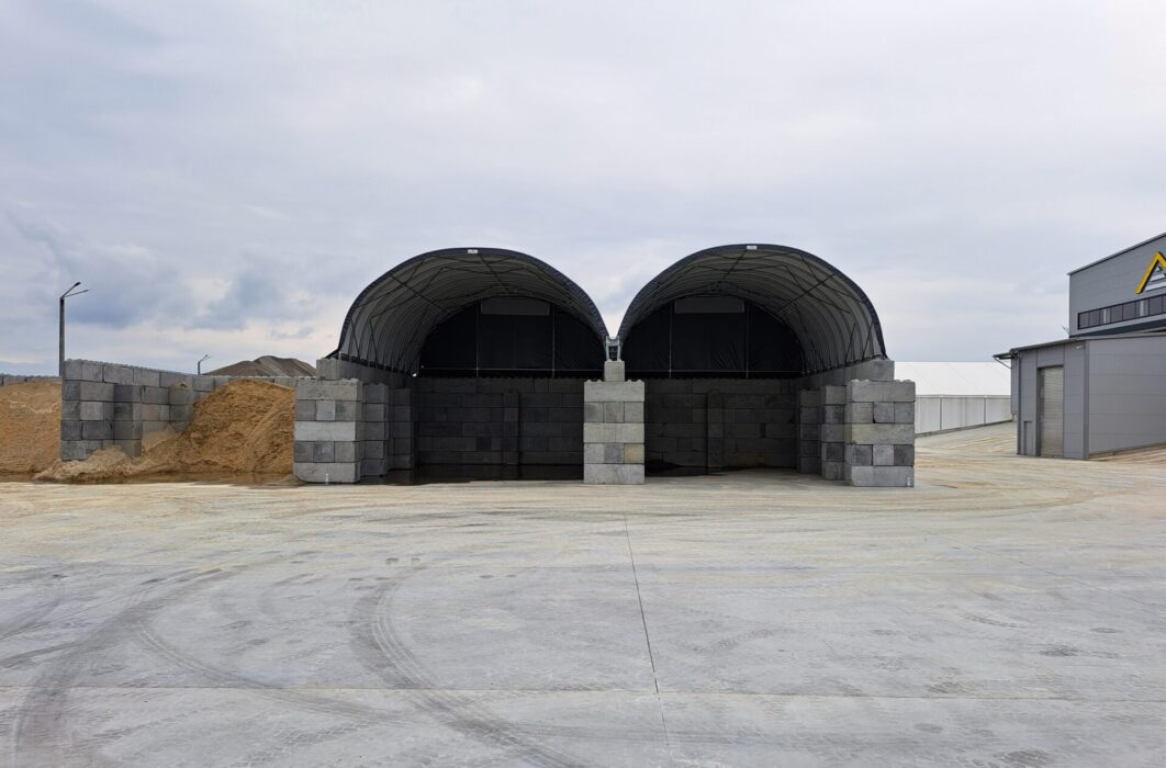 Dwie duże hale namiotowe z betonowymi ścianami, przeznaczone do składowania piasku i kruszywa. Konstrukcje łukowe chronią materiały budowlane przed deszczem i wiatrem, zapewniając bezpieczne warunki magazynowania. Idealne rozwiązanie dla firm z branży budowlanej i drogowej.