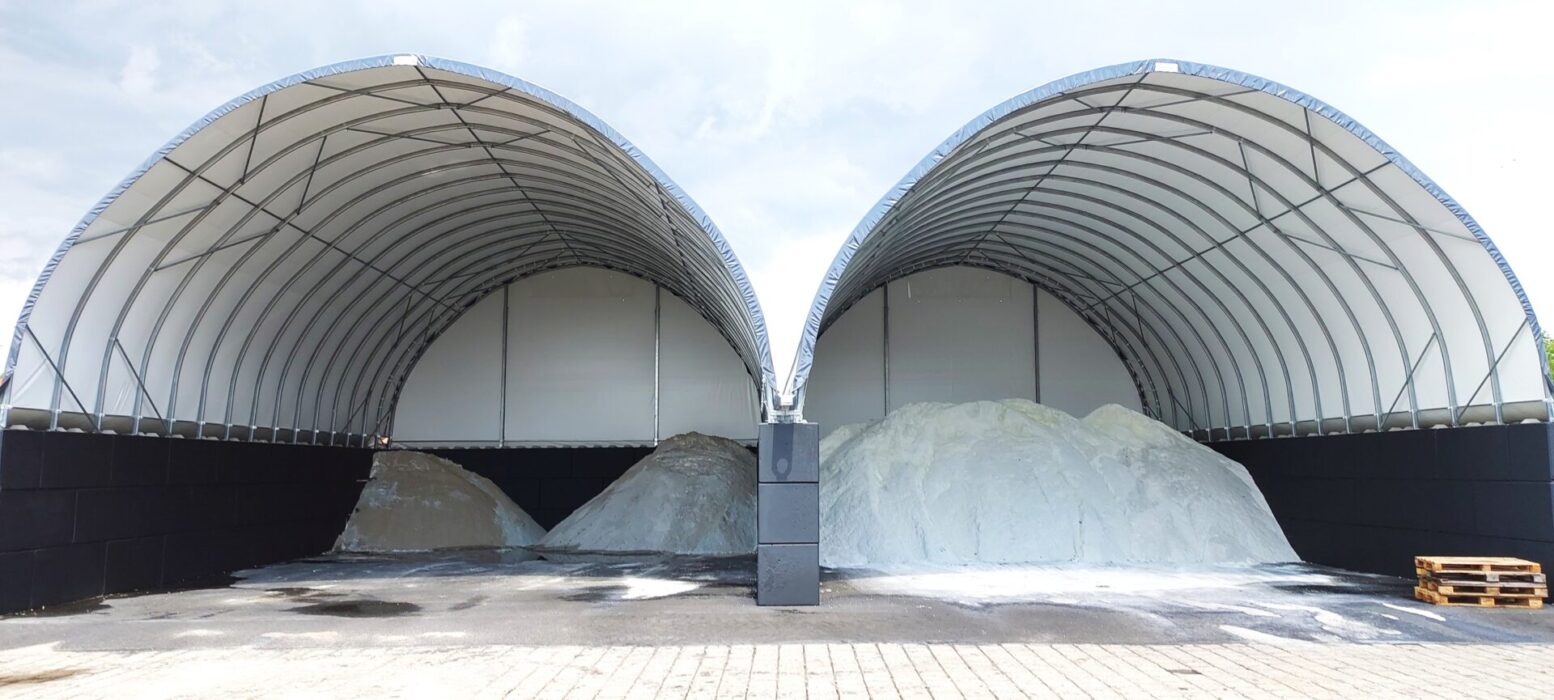 Na zdjęciu widoczne są dwie połączone hale łukowe przeznaczone do składowania soli drogowej i piasku. Wewnątrz znajdują się duże pryzmy materiału o różnych odcieniach, a konstrukcja opiera się na stalowym szkielecie z jasnym poszyciem namiotowym i wysokimi ścianami z betonowych bloków. Plac przed halą jest utwardzony, co ułatwia obsługę i transport surowców.. Oddzielone są hałdy piasku oraz soli drogowej.