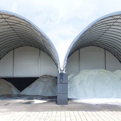 Na zdjęciu widoczne są dwie połączone hale łukowe przeznaczone do składowania soli drogowej i piasku. Wewnątrz znajdują się duże pryzmy materiału o różnych odcieniach, a konstrukcja opiera się na stalowym szkielecie z jasnym poszyciem namiotowym i wysokimi ścianami z betonowych bloków. Plac przed halą jest utwardzony, co ułatwia obsługę i transport surowców.. Oddzielone są hałdy piasku oraz soli drogowej.