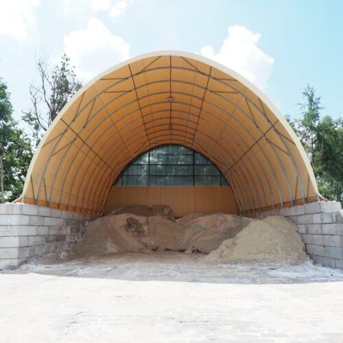 Na zdjęciu znajduje się hala łukowa przeznaczona do składowania soli i piasku drogowego. Wewnątrz widoczne są duże pryzmy materiału, a konstrukcja opiera się na ścianach z betonowych bloków i żółtym poszyciu namiotowym. Hala zlokalizowana jest na utwardzonym placu, w otoczeniu drzew.