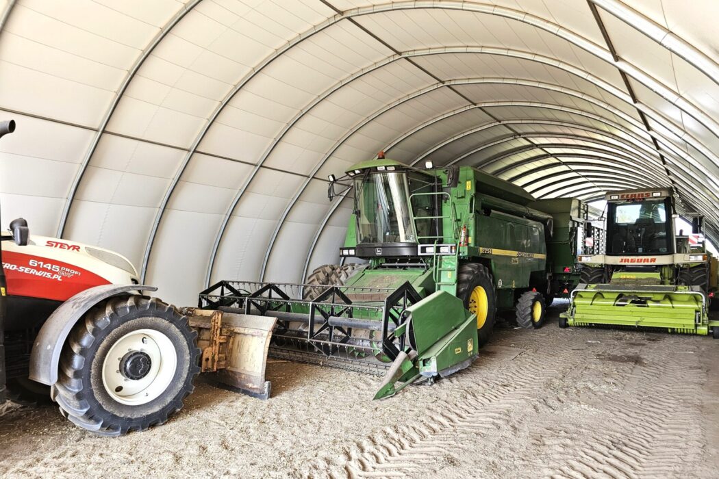 Na zdjęciu znajduje się hala namiotowa pełniąca funkcję garażu na kombajny. Wewnątrz zaparkowane są kombajny i traktor, ustawione w rzędzie w przestronnej konstrukcji łukowej. Hala chroni pojazdy przed deszczem, słońcem i innymi warunkami atmosferycznymi.