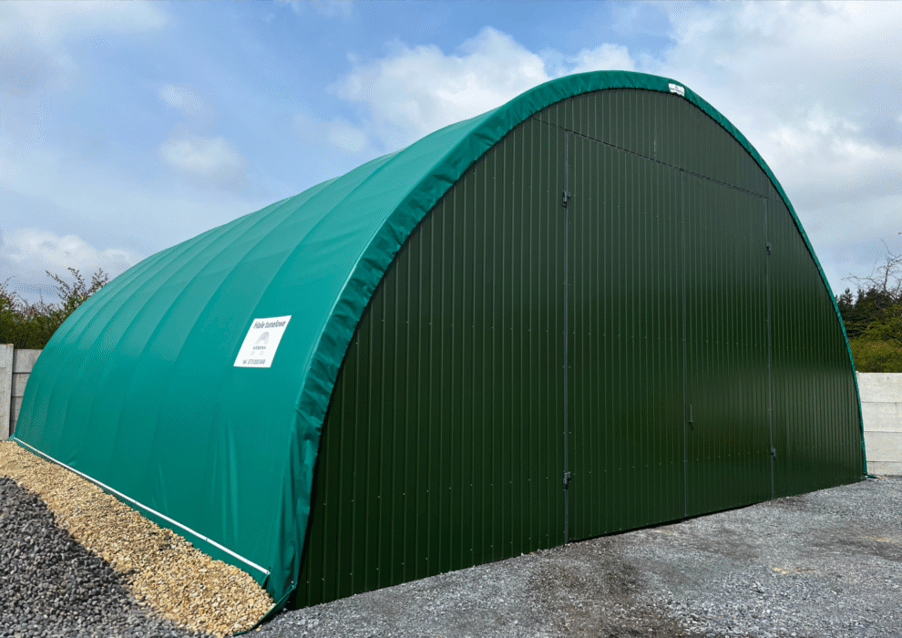 Zielona hala łukowa z zamkniętymi dwuskrzydłowymi drzwiami frontowymi, ustawiona na utwardzonym podłożu z tłucznia i otoczona betonowym ogrodzeniem.