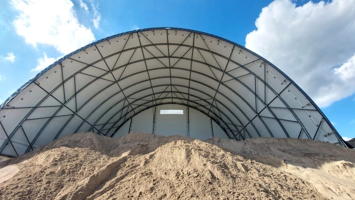 Na zdjęciu znajduje się hala łukowa przeznaczona do składowania kruszyw. Wewnątrz widoczne są duże pryzmy piasku, a konstrukcja opiera się na stalowym szkielecie pokrytym jasnym poszyciem namiotowym. Hala chroni materiał przed deszczem i wiatrem, zapewniając odpowiednie warunki magazynowania.