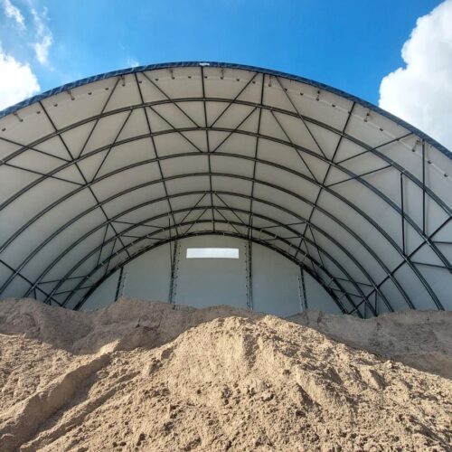 Na zdjęciu znajduje się hala łukowa przeznaczona do składowania kruszyw. Wewnątrz widoczne są duże pryzmy piasku, a konstrukcja opiera się na stalowym szkielecie pokrytym jasnym poszyciem namiotowym. Hala chroni materiał przed deszczem i wiatrem, zapewniając odpowiednie warunki magazynowania.