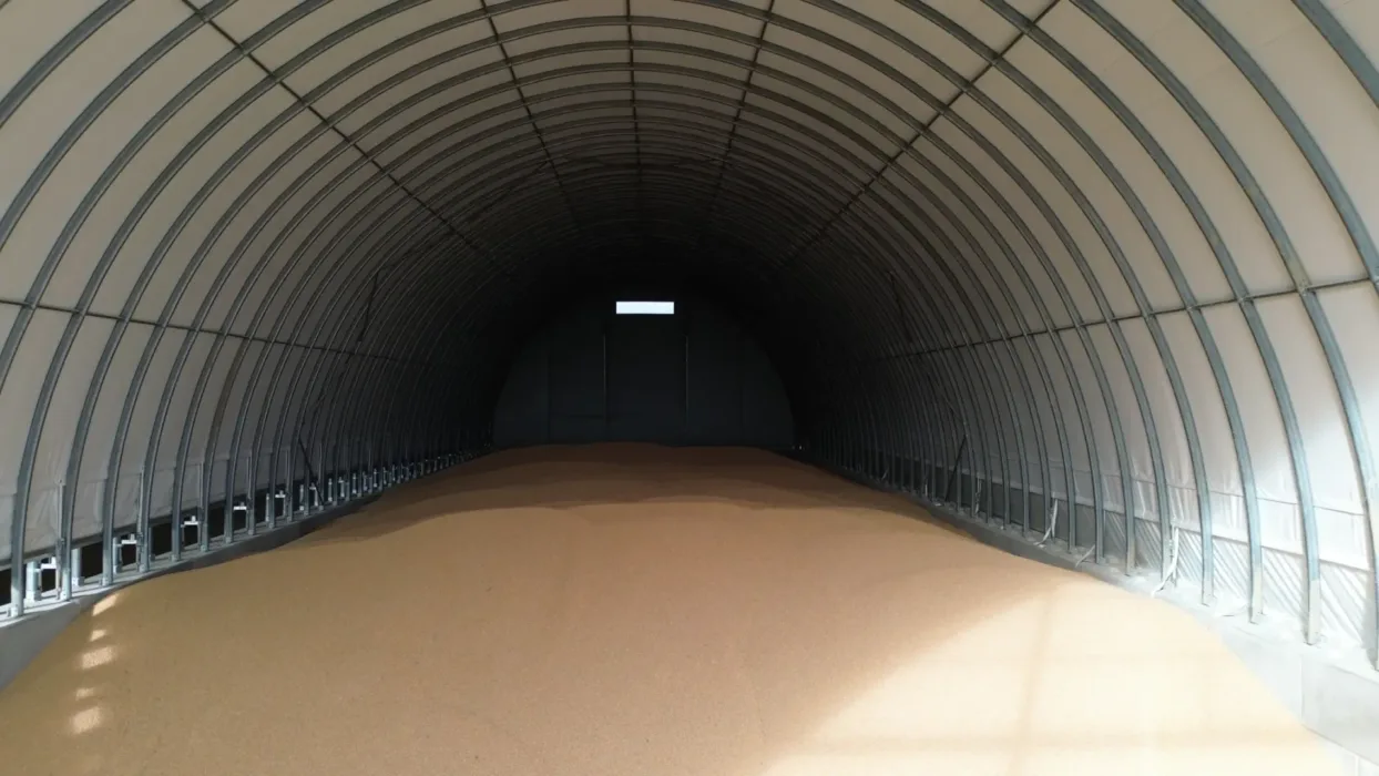 Wnętrze pustej hali magazynowej na zboże z charakterystyczną łukową konstrukcją stalową. Hala ma betonową podłogę i białe ściany boczne, z widocznym oświetleniem w głębi budynku.