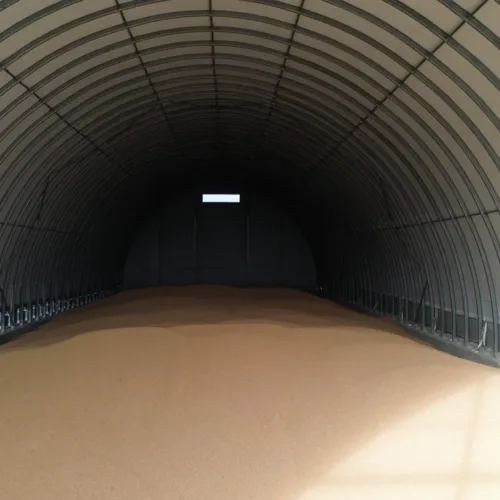 Wnętrze pustej hali magazynowej na zboże z charakterystyczną łukową konstrukcją stalową. Hala ma betonową podłogę i białe ściany boczne, z widocznym oświetleniem w głębi budynku.