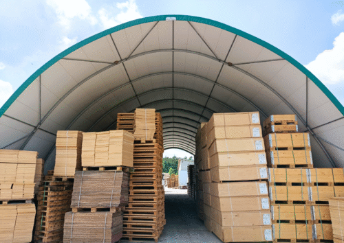 Hala magazynowa o łukowej konstrukcji z jasnym poszyciem, wypełniona paletami z drewnianymi płytami i skrzyniami transportowymi. Obiekt zapewnia zadaszoną i przestronną przestrzeń do składowania materiałów przemysłowych i produkcyjnych.