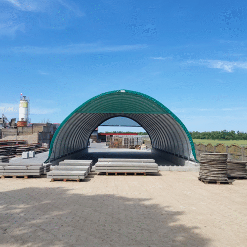 Hala namiotowa o łukowej konstrukcji z zielonym poszyciem, wykorzystywana na placu przemysłowym do składowania prefabrykatów betonowych. Wokół obiektu widoczne są palety z elementami budowlanymi, co podkreśla jej zastosowanie w branży produkcyjno-montażowej.