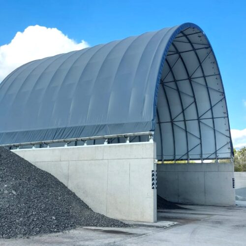 Hala namiotowa z wysokimi betonowymi ścianami, przeznaczona do składowania destruktu asfaltowego i kruszywa. Przed wejściem do hali widoczna jest duża pryzma ciemnego materiału, a w tle mniejsza hałda jasnego kruszywa. Konstrukcja znajduje się na otwartym placu przemysłowym pod gołym niebem.