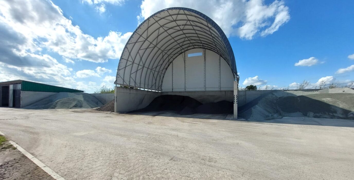 Duża hala namiotowa z betonowymi ścianami, służąca do przechowywania destruktu asfaltowego i kruszywa. Wewnątrz widoczne są ciemne hałdy materiału, a po bokach jasne pryzmy żwiru i kamienia. Konstrukcja stoi na utwardzonym placu pod otwartym niebem.