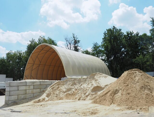 Na zdjęciu widoczna jest hala łukowa przeznaczona do składowania kruszyw. Konstrukcja opiera się na ścianach z betonowych bloków i przykryta jest jasnym poszyciem namiotowym. Przed halą usypane są duże pryzmy piasku i kruszywa, a całość znajduje się na otwartym placu w otoczeniu drzew.