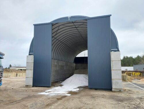 Na zdjęciu znajduje się hala łukowa z wysokimi betonowymi ścianami i metalowymi drzwiami przesuwnymi, przeznaczona do składowania soli drogowej i piasku. Wewnątrz widoczne są pryzmy materiału, a utwardzony plac przed halą umożliwia swobodny dojazd pojazdów ciężarowych. Konstrukcja zapewnia ochronę surowców przed czynnikami atmosferycznymi.