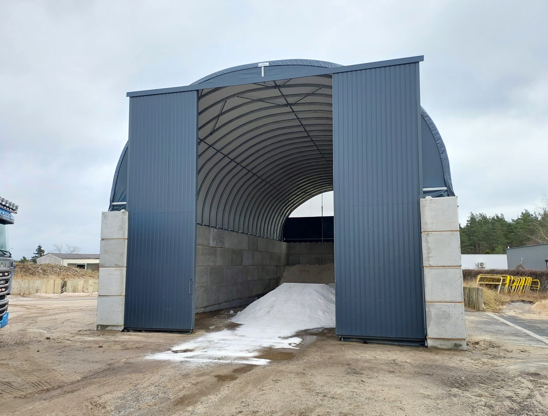 Na zdjęciu znajduje się hala łukowa z wysokimi betonowymi ścianami i metalowymi drzwiami przesuwnymi, przeznaczona do składowania soli drogowej i piasku. Wewnątrz widoczne są pryzmy materiału, a utwardzony plac przed halą umożliwia swobodny dojazd pojazdów ciężarowych. Konstrukcja zapewnia ochronę surowców przed czynnikami atmosferycznymi.