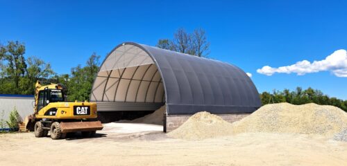 Na zdjęciu znajduje się hala łukowa przeznaczona do składowania żwiru, z wysokimi ścianami z bloczków betonowych i ciemnym poszyciem namiotowym. Przed halą widoczne są duże pryzmy żwiru oraz żółta koparka marki Caterpillar. Całość zlokalizowana jest na placu budowlanym otoczonym zielenią.