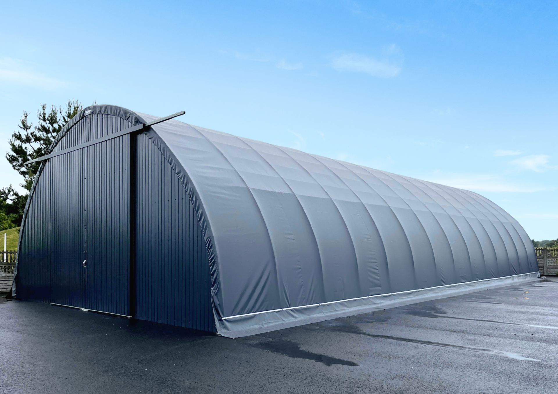 Hala namiotowa o łukowej konstrukcji z szarym poszyciem i dużymi bramami wjazdowymi, ustawiona na utwardzonym placu asfaltowym. Obiekt stanowi uniwersalną przestrzeń magazynową, przystosowaną do przechowywania towarów i sprzętu w różnych warunkach pogodowych.