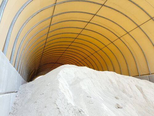 Wnętrze hali łukowej wypełnione jest dużą pryzmą kredy. Ściany wykonane z betonowych bloków oraz żółte poszycie namiotowe chronią materiał przed czynnikami atmosferycznymi. Konstrukcja zapewnia bezpieczne i wydajne przechowywanie surowca.