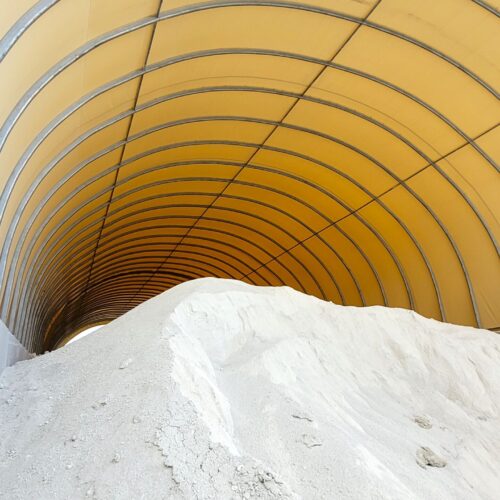 Wnętrze hali łukowej wypełnione jest dużą pryzmą kredy. Ściany wykonane z betonowych bloków oraz żółte poszycie namiotowe chronią materiał przed czynnikami atmosferycznymi. Konstrukcja zapewnia bezpieczne i wydajne przechowywanie surowca.