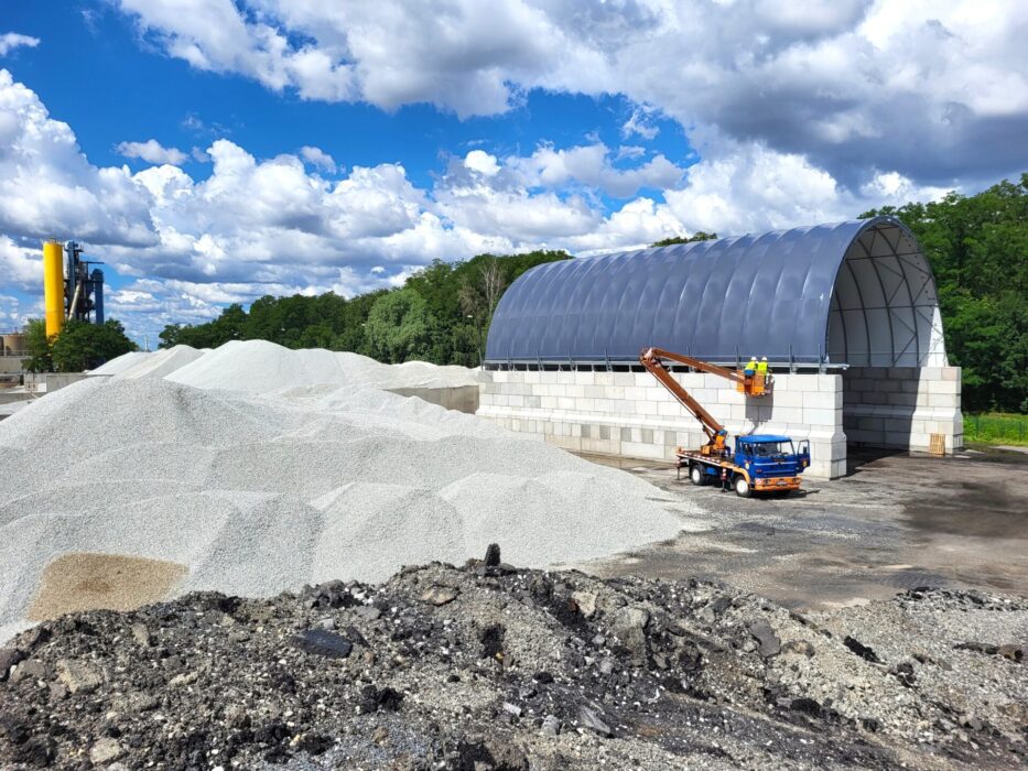 Hala łukowa z betonowymi ścianami bocznymi wykorzystywana do składowania destruktu asfaltowego. Obok hali widać duże pryzmy jasnego kruszywa budowlanego oraz pracowników na podnośniku montujących elementy konstrukcji. Całość znajduje się na otwartej przestrzeni przemysłowej.