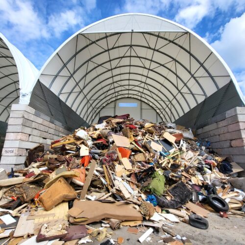 Na zdjęciu widać halę namiotową wykorzystywaną do składowania odpadów wielkogabarytowych. Wewnątrz znajduje się duża sterta różnorodnych odpadów, m.in. fragmenty mebli, materace, deski, elementy tapicerki, plastikowe przedmioty i opony. Konstrukcja hali opiera się na stalowym szkielecie i betonowych ścianach oporowych, a dach pokryty jest jasną membraną chroniącą zawartość przed warunkami atmosferycznymi.