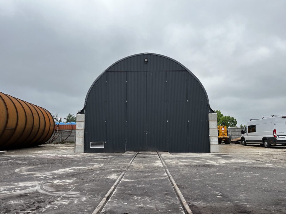 Czarna hala namiotowa o konstrukcji łukowej, z dużymi, zamkniętymi drzwiami frontowymi i betonowymi elementami bocznymi, stojąca na placu przemysłowym.