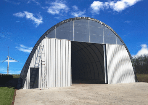 Hala namiotowa z metalową elewacją i dużymi przesuwnymi drzwiami, stojąca na utwardzonym placu, z drabiną opartą o ścianę i widoczną w tle turbiną wiatrową