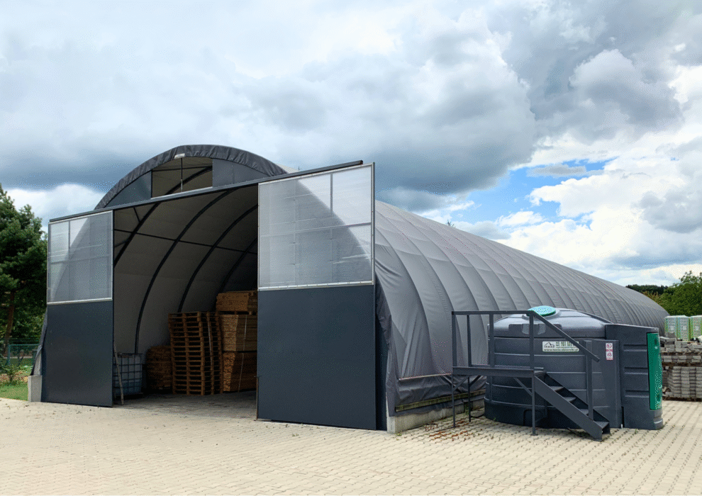 Hala łukowa z otwartymi drzwiami przesuwnymi, wypełniona drewnianymi paletami i deskami. Obok hali stoi duży zbiornik zewnętrzny z metalowymi schodkami. Na placu widać kostkę brukową i elementy składowe, takie jak bloczki lub płyty.