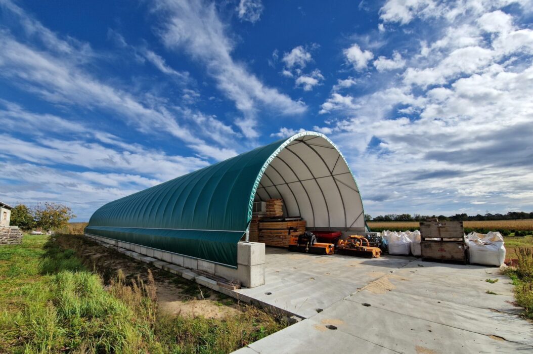 Hala łukowa z zielonym poszyciem zewnętrznym, usytuowana na betonowym fundamencie na otwartym terenie. Wewnątrz widoczne są ułożone palety i maszyny rolnicze, a obok hali stoją big bagi i skrzynie.