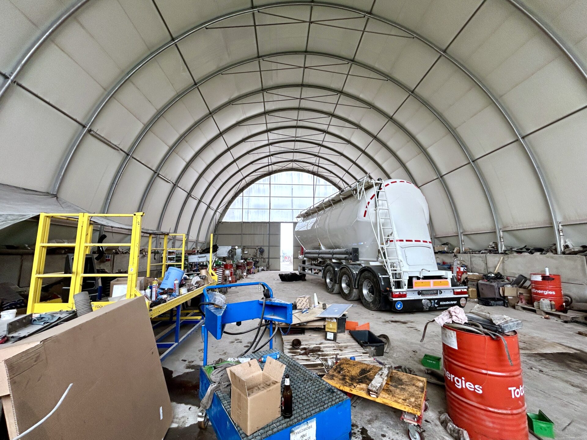 Wnętrze hali łukowej z serwisowaną naczepą ciężarową i stanowiskami warsztatowymi. Po lewej stronie widoczny jest żółty podest serwisowy i różnorodne narzędzia, a po prawej beczki olejowe oraz części mechaniczne.