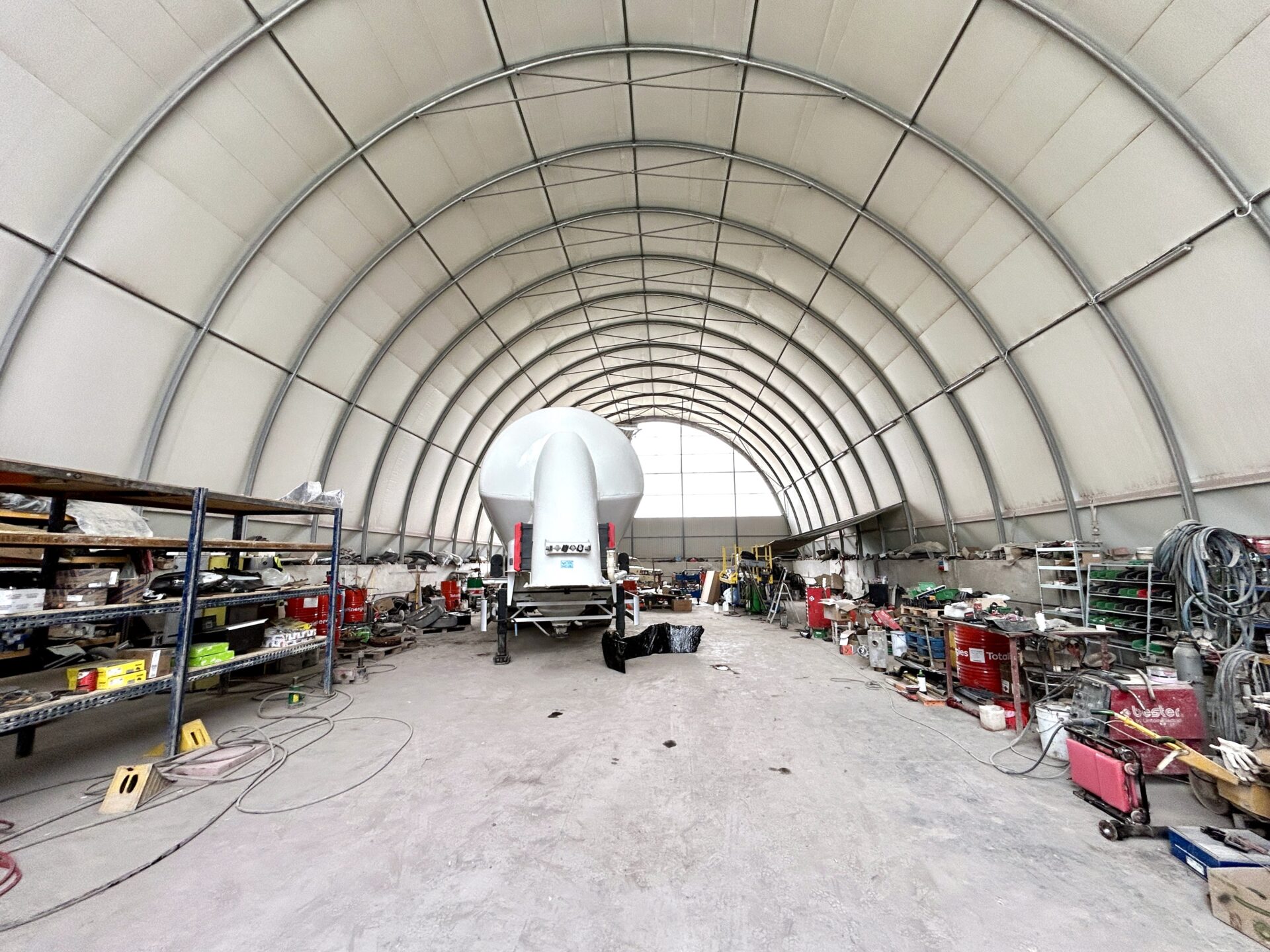 Wnętrze łukowej hali warsztatowej z naczepą do transportu sypkich materiałów, otoczoną licznymi regałami, narzędziami i sprzętem serwisowym.