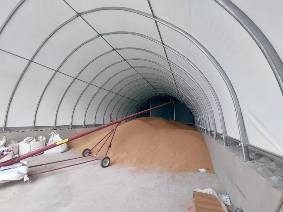 Hala magazynowa podczas procesu napełniania zbożem za pomocą systemu transportowego. Widoczny jest duży stożek ziarna oraz czerwone przenośniki taśmowe, a także worki big-bag po lewej stronie.