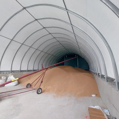 Hala magazynowa podczas procesu napełniania zbożem za pomocą systemu transportowego. Widoczny jest duży stożek ziarna oraz czerwone przenośniki taśmowe, a także worki big-bag po lewej stronie.