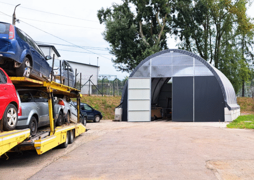Widok zewnętrzny hali łukowej z blachy i tworzywa, z częściowo otwartymi drzwiami przesuwnymi. Przed halą stoi laweta z wieloma samochodami osobowymi, co sugeruje, że obiekt pełni funkcję warsztatu, punktu demontażu lub magazynu pojazdów używanych.
