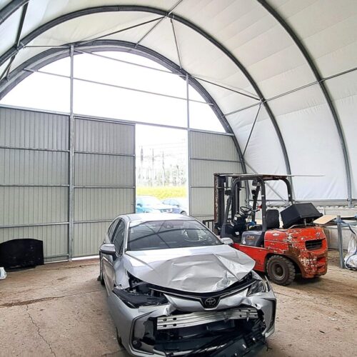 Hala namiotowa pełniąca funkcję warsztatu, z betonowymi murkami oporowymi i metalowym stelażem konstrukcyjnym. Wewnątrz znajduje się uszkodzony samochód osobowy, wózek widłowy oraz różne narzędzia i elementy wyposażenia warsztatowego. Duża brama wjazdowa zapewnia łatwy dostęp do wnętrza obiektu.