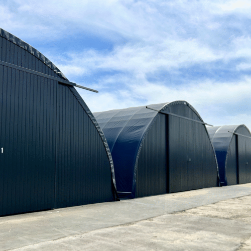 Kompleks hal namiotowych o łukowej konstrukcji z ciemnym poszyciem i dużymi bramami wjazdowymi. Obiekty ustawione w rzędzie tworzą funkcjonalną przestrzeń magazynową, idealną do przechowywania towarów i sprzętu.