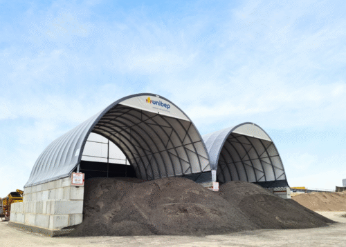 Dwie hale łukowe z logo UNIBEP wypełnione pryzmami destruktu asfaltowego na placu budowy