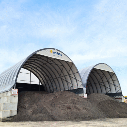 Dwie hale łukowe z logo UNIBEP wypełnione pryzmami destruktu asfaltowego na placu budowy