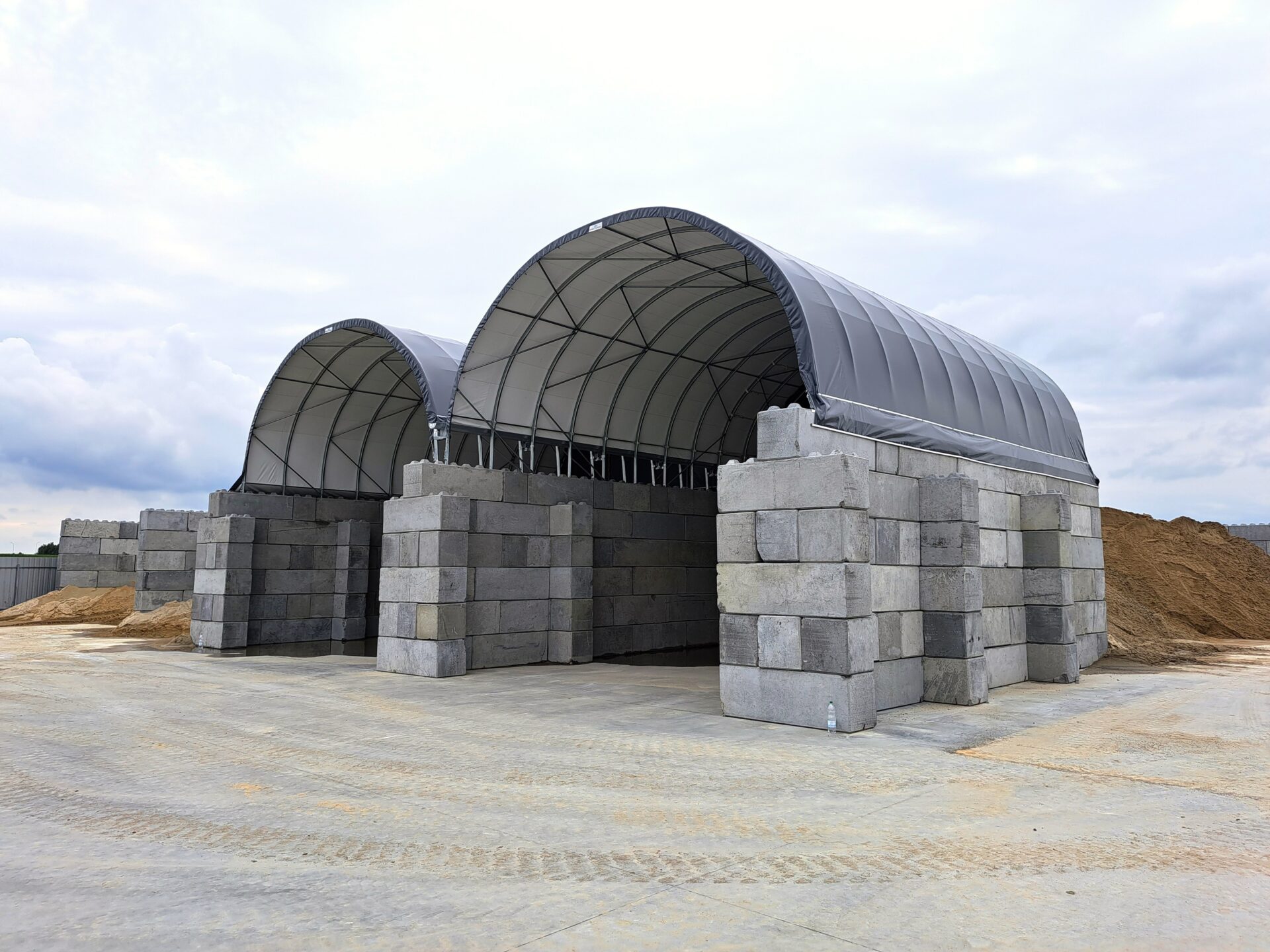 Dwie hale namiotowe z solidnymi betonowymi ścianami, wykorzystywane do przechowywania piasku i kruszyw. Konstrukcja łukowa z wytrzymałym poszyciem chroni materiały przed opadami i wiatrem. Rozwiązanie idealne dla składów budowlanych i firm drogowych.