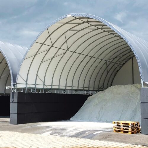 Na zdjęciu widoczne są dwie hale łukowe przeznaczone do składowania soli drogowej. W jednej z hal znajduje się duża pryzma białego materiału, a druga jest pusta. Konstrukcje oparte są na ścianach z betonowych bloków i przykryte jasnym poszyciem namiotowym.