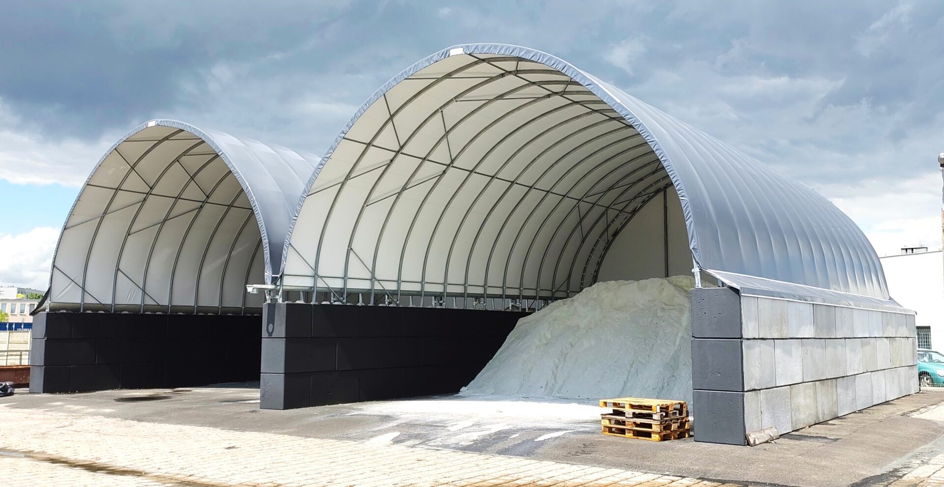 Na zdjęciu widoczne są dwie hale łukowe przeznaczone do składowania soli drogowej. W jednej z hal znajduje się duża pryzma białego materiału, a druga jest pusta. Konstrukcje oparte są na ścianach z betonowych bloków i przykryte jasnym poszyciem namiotowym.